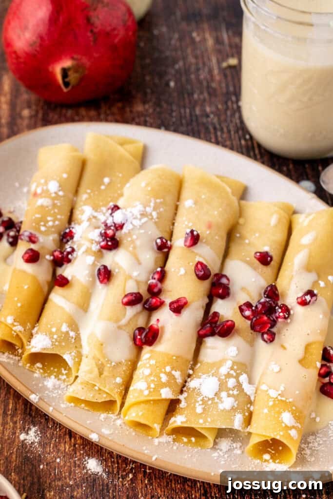 Close-up of rolled eggnog crepes drizzled with eggnog sauce, sprinkled with powdered sugar, and topped with bright pomegranate arils, highlighting the festive presentation.