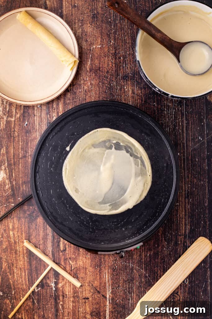 Finished eggnog crepe cooking on the pan with one rolled crepe on a plate in the background, ready for serving, showcasing progress.