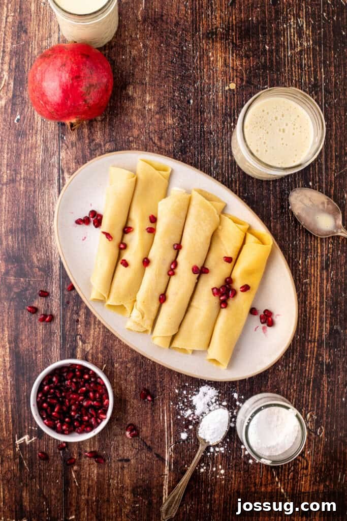 A plate of plain rolled eggnog crêpes on a rustic wood table, surrounded by pomegranate seeds, a jar of eggnog sauce, and a spoon with powdered sugar, awaiting garnishing.