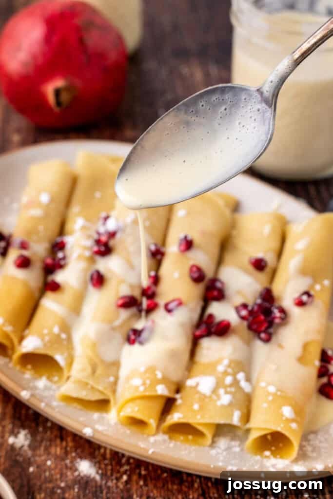 A close-up of a spoon drizzling creamy eggnog sauce over rolled crêpes topped with pomegranate seeds and a dusting of powdered sugar, demonstrating the final flourish.