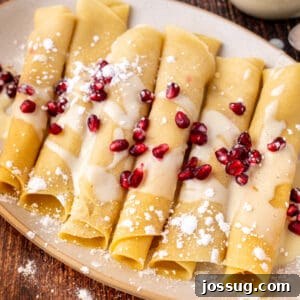 Square crop of eggnog crepes drizzled with eggnog sauce and topped with pomegranate arils, served on a beige plate over rustic wood, highlighting the festive dish.