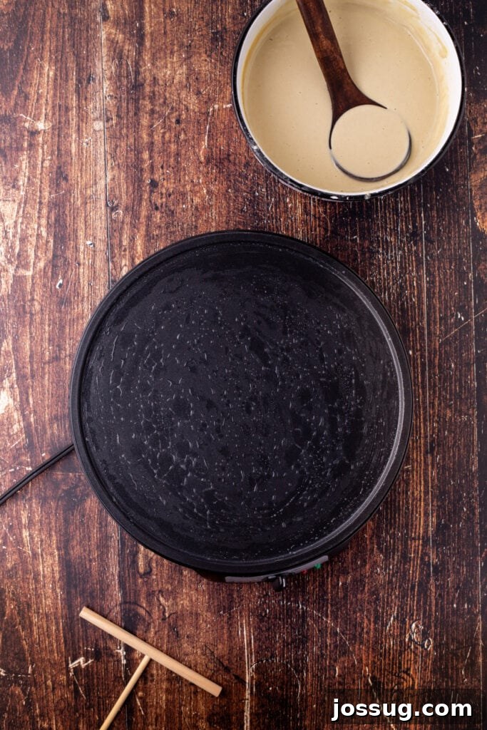 Crepe pan preheated and ready for cooking, with a bowl of batter and a wooden ladle nearby on a rustic wood table, setting the scene for crepe making.