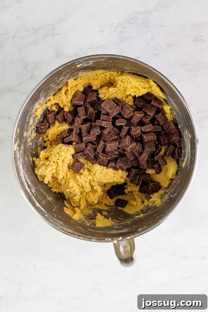 A pile of rich chocolate chunks sitting on top of the thoroughly mixed cookie dough in a mixing bowl, waiting to be folded in.