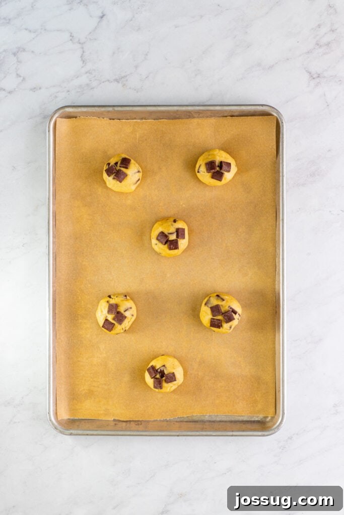 Cookie dough balls with additional chocolate chunks pressed onto their tops, arranged on a parchment-lined baking sheet for a professional finish.