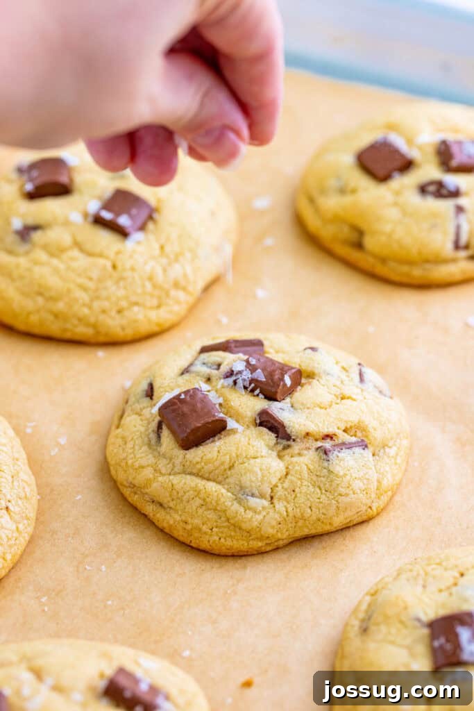A hand gently sprinkling flaky sea salt over warm, freshly baked chocolate chunk cookies on a baking sheet, highlighting the finishing touch.