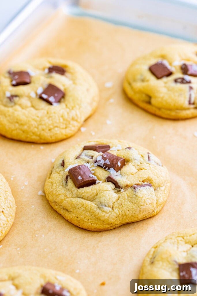 A close-up shot of warm chocolate chunk cookies with melted chocolate and shimmering flaky sea salt, cooling on a parchment-lined baking sheet.