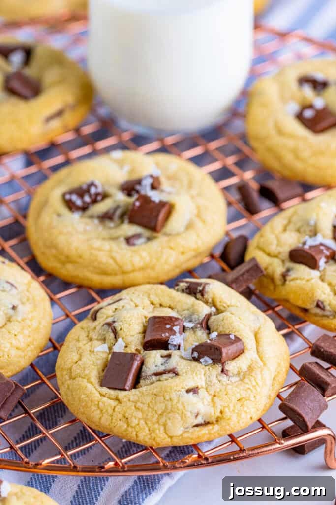 Delicious chocolate chunk cookies, topped with flaky salt, cooling on a copper wire rack with a blurred glass of milk in the background, ready to be enjoyed.