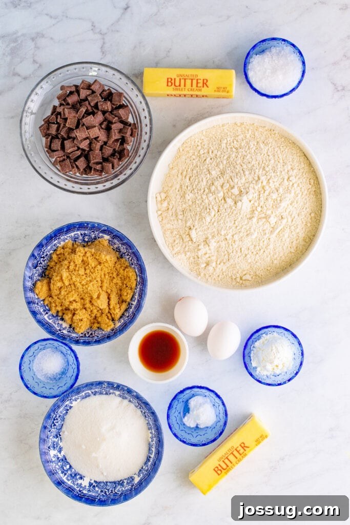 Overhead view of all cookie ingredients neatly arranged in separate bowls, including flour, two types of sugar, softened butter, large eggs, vanilla extract, and a large bowl of semi-sweet chocolate chunks.