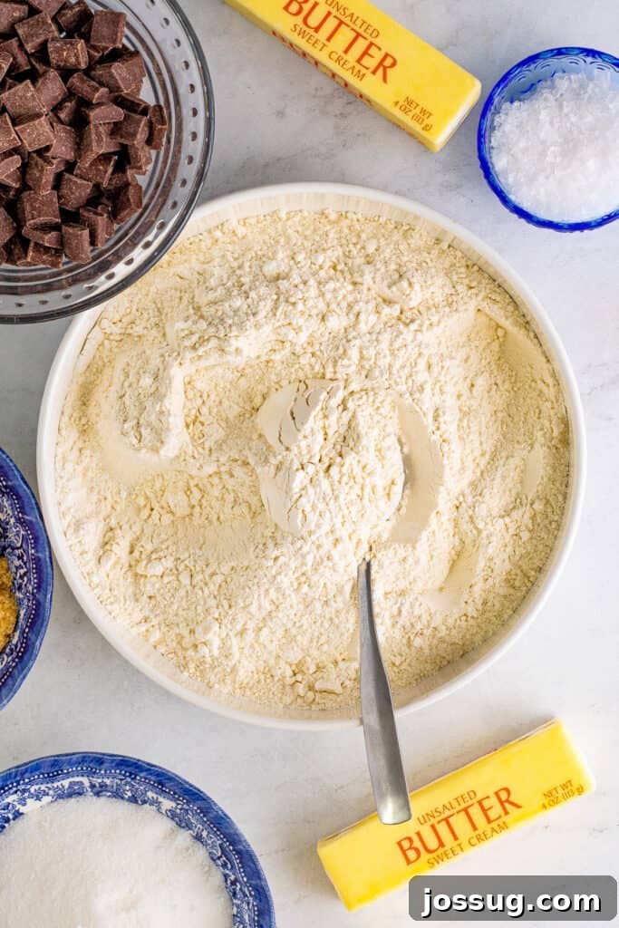 A bowl of flour mixture with a spoon, surrounded by butter, sugars, eggs, vanilla, and chocolate chunks - all necessary ingredients for the cookie dough.