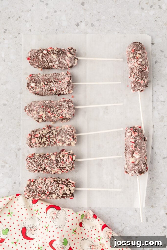 Peppermint Cloud Wands 11 A collection of beautifully finished peppermint marshmallow wands, coated in chocolate and sparkling with crushed peppermint pieces, neatly arranged on parchment paper to cool and fully set.