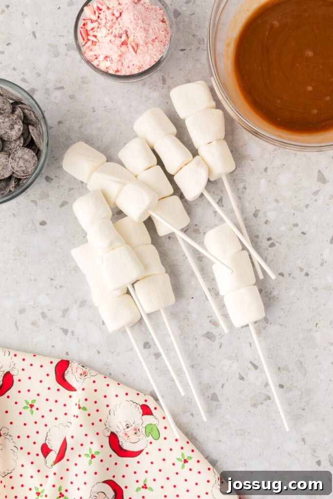 Peppermint Cloud Wands 5 Several lollipop sticks, each meticulously holding three fluffy marshmallows, neatly arranged on a countertop beside a bowl of melted caramel and another of vibrant crushed peppermint, showcasing the assembly stage.