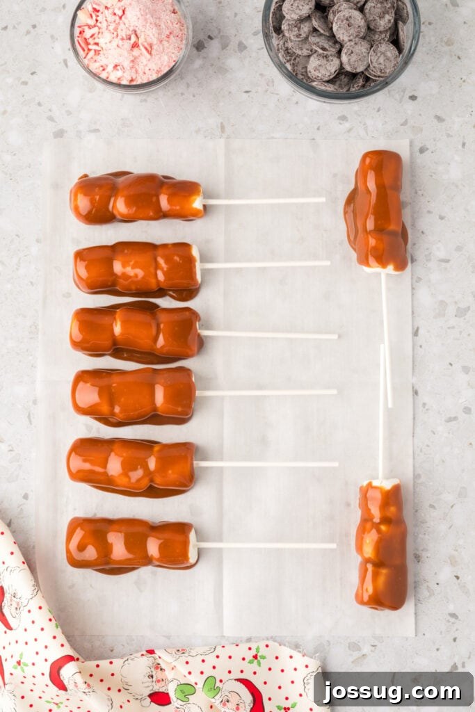 Peppermint Cloud Wands 6 Marshmallow wands, now beautifully coated in a glossy caramel layer, resting on a sheet of parchment paper to set, with bowls of dark chocolate wafers and crushed peppermint waiting nearby for the next step.