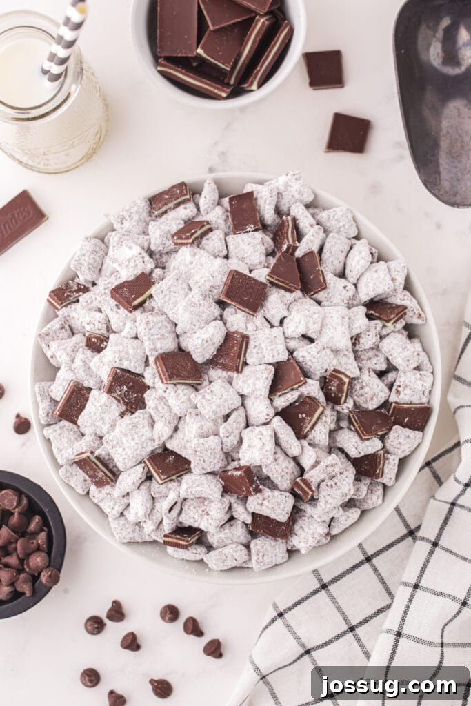 Bowl of Thin Mint Muddy Buddies topped with chopped Andes mints, next to a bottle of milk, chocolate chips, and a striped cloth.