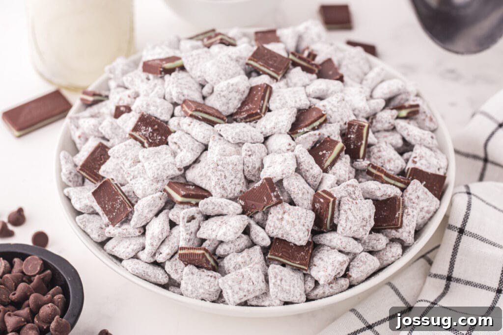 Bowl of Thin Mint Muddy Buddies topped with chopped Andes mints, with chocolate chips and a glass bottle of milk in the background.