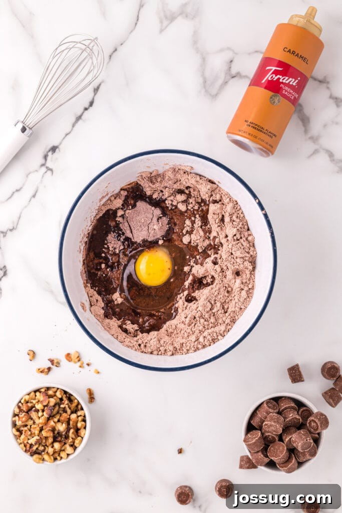 Mixing bowl with brownie mix, egg, water, and oil before being stirred, surrounded by walnuts, Rolos, caramel sauce, and a whisk on a marble surface.