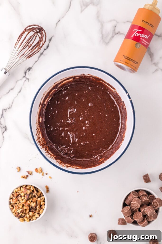 Smooth brownie batter mixed in a white bowl with a whisk and caramel sauce bottle nearby, with walnuts and Rolos scattered around.