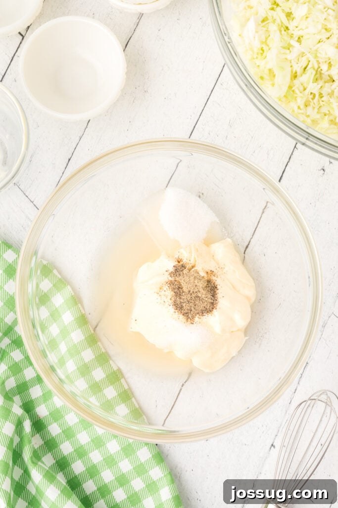 Glass bowl with coleslaw dressing ingredients before mixing, next to a whisk and green kitchen towel.