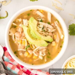 Square crop of a bowl of White Chicken Chili with avocado slices and cheese on top, served on a red and aqua towel with cilantro garnish.