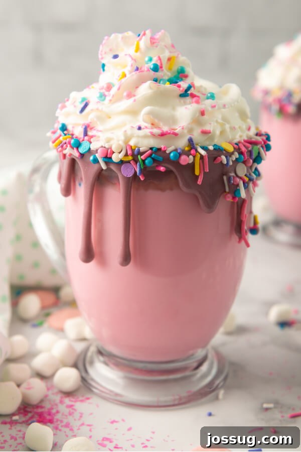 Rainbow Dream Cocoa 2 A vibrant glass mug filled with pink Unicorn Hot Chocolate, topped with a mountain of whipped cream, an abundance of colorful sprinkles, and various pastel marshmallows.