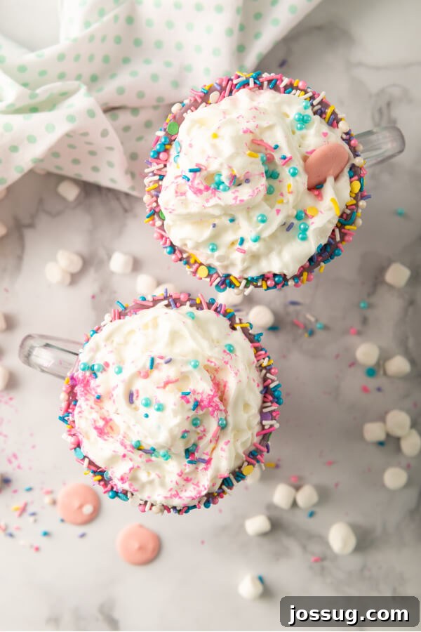 Rainbow Dream Cocoa 7 A top-down view of two beautifully decorated mugs of Unicorn Hot Chocolate, brimming with whipped cream, colorful sprinkles, and pastel marshmallows, on a rustic wooden surface.