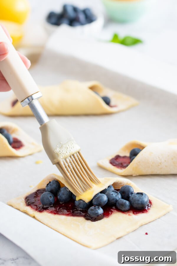 Bright Lemon Blueberry Danish Delight 6 The initial step of folding a corner of the puff pastry over the blueberry filling, brushed with egg wash