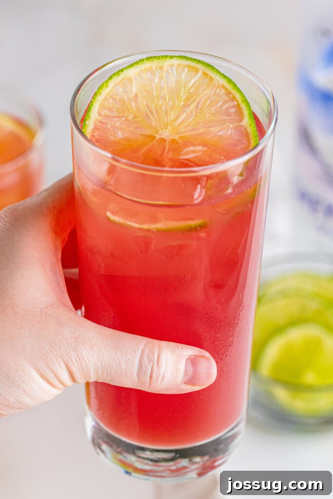 Coastal Sunset Sipper 4 A hand holding a tall glass with a Bay Breeze cocktail, garnished with a vibrant lime wheel, against a soft blurred background.