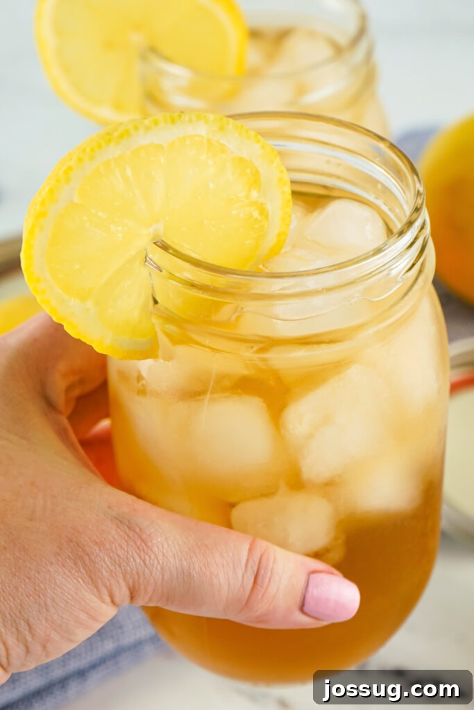 Hand holding a glass mason jar filled with a Spiked Arnold Palmer cocktail, garnished with a lemon slice, against a bright outdoor background.