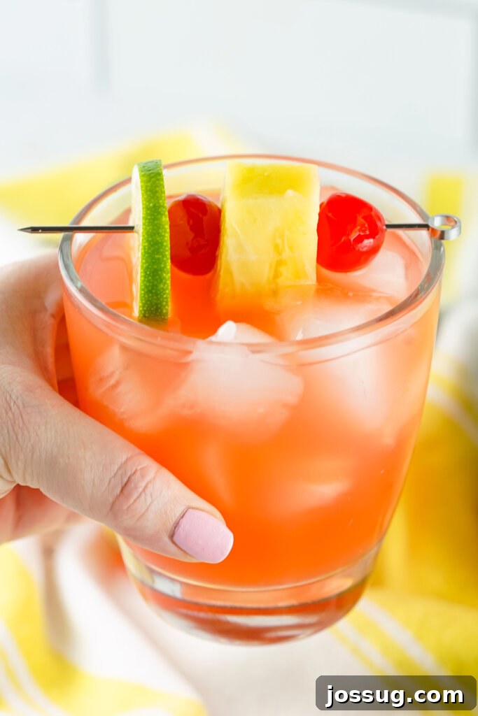 Tropical Rum Punch 4 A person holding a beautifully garnished glass of Rum Punch Cocktail, featuring a lime wedge and a maraschino cherry, against a blurred tropical beach background, evoking a sense of relaxation.