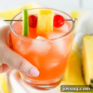 Hand holding a vibrant Rum Punch Cocktail garnished with a lime wedge and cherry.