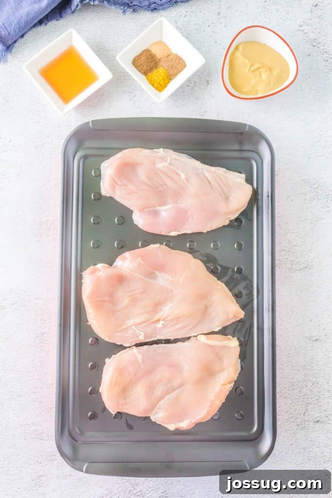 Raw chicken breasts meticulously arranged on a marinade tray, surrounded by small bowls of various seasonings, a bottle of honey, and a jar of Dijon mustard, illustrating the preparation stage.