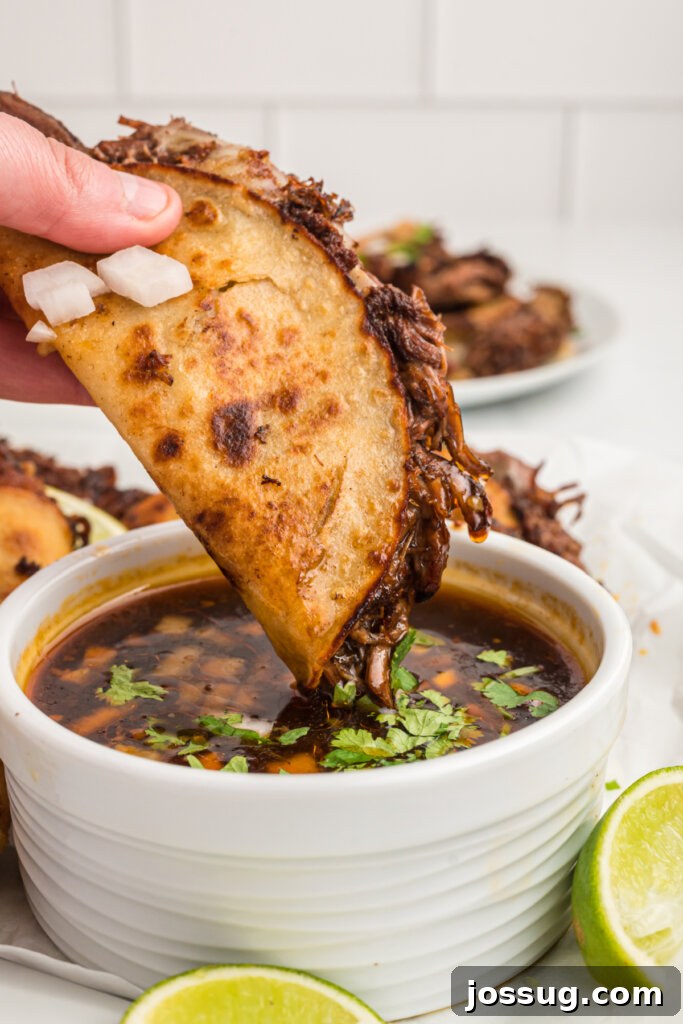 Hand dipping a birria taco into rich, red consommé