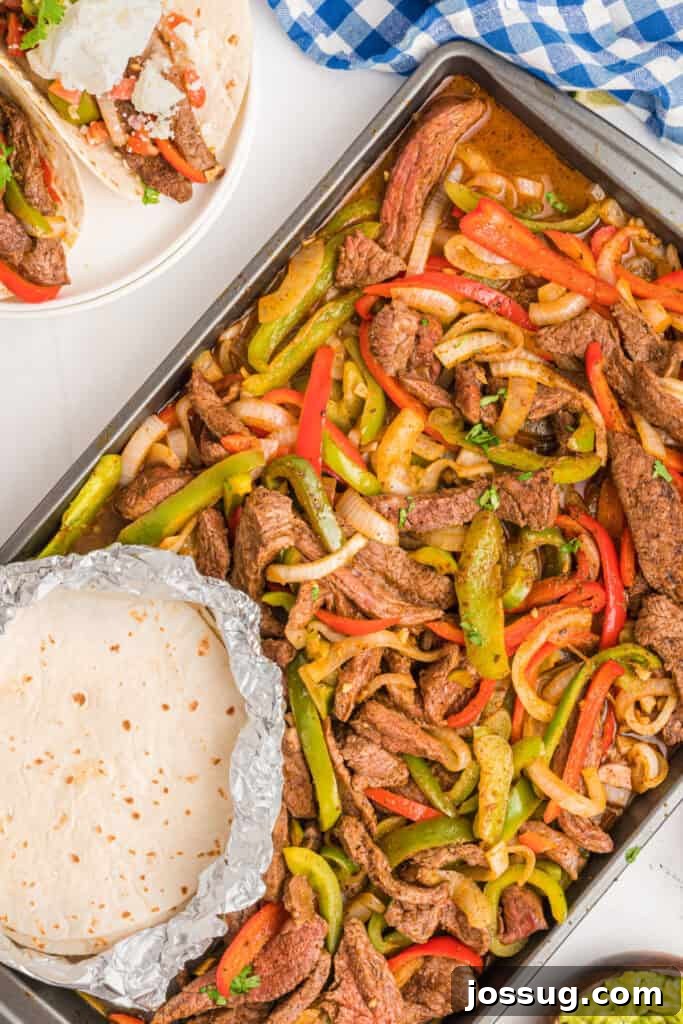 Full sheet pan of cooked steak fajitas with tortillas in foil and a partial plate of tacos in the corner.