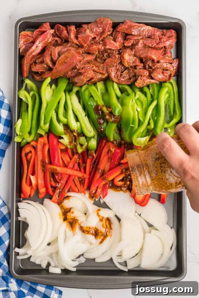 Raw steak and sliced peppers and onions arranged on a sheet pan with marinade being poured over.