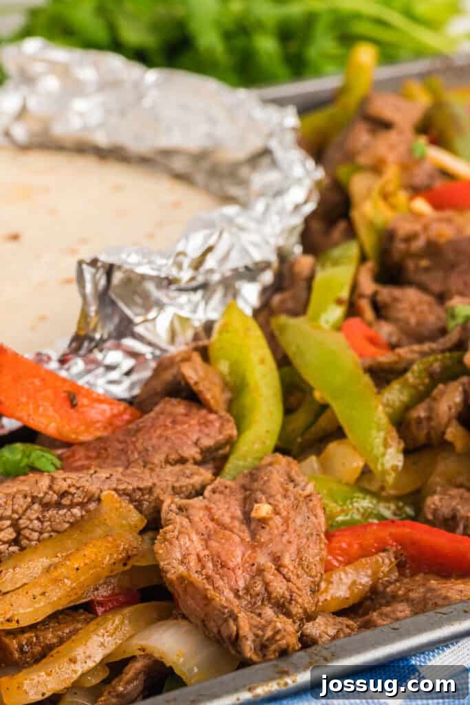 Sheet pan of cooked steak fajitas with foil-wrapped tortillas in the corner and fresh herbs in the background.