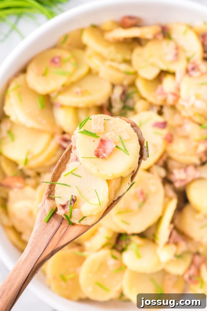 A wooden spoon lifting a serving of German potato salad from a larger serving bowl, highlighting the sliced potatoes and bacon bits.