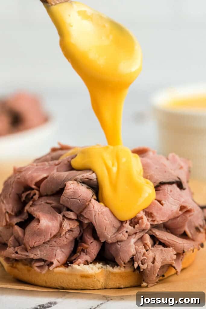 Delicious, golden cheddar cheese sauce being generously drizzled from a spoon onto a perfectly assembled roast beef sandwich.