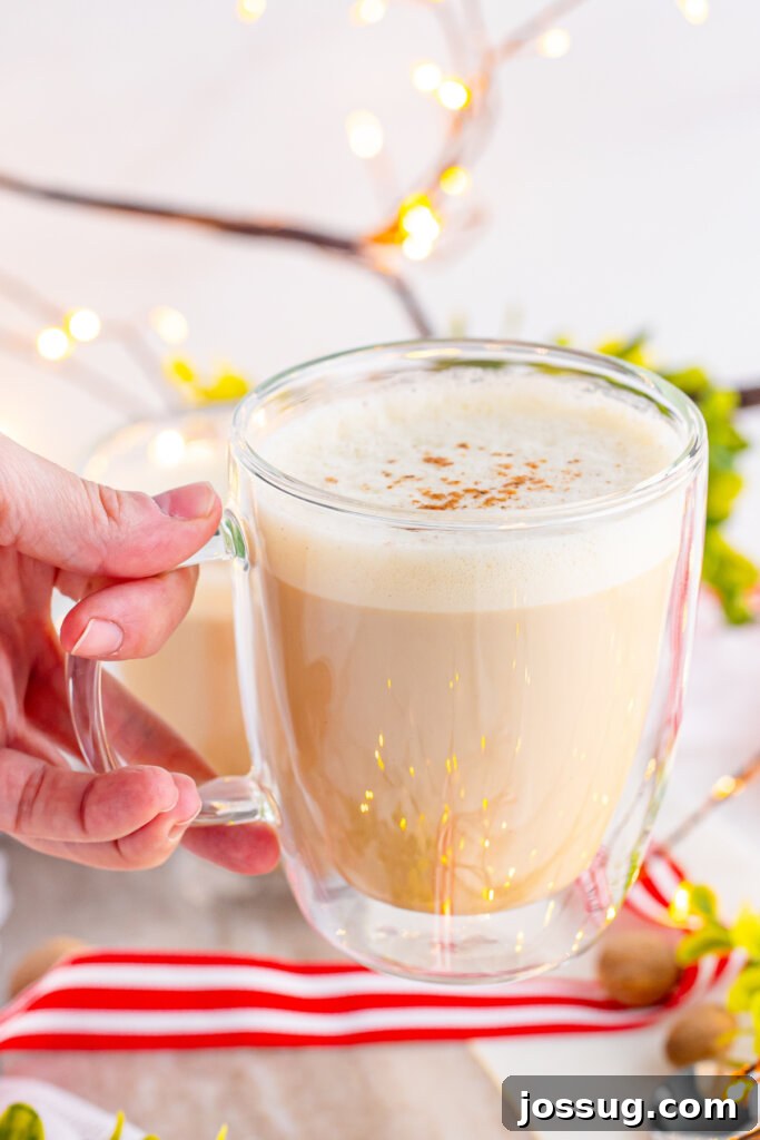 Yuletide Velvet Latte 5 A hand holding a clear mug of eggnog latte, with a cozy, festive background, emphasizing the warmth and comfort of the drink.