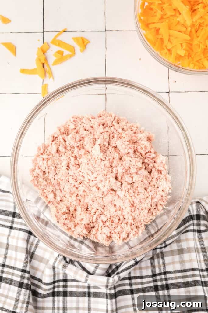Cheesy Sausage Balls 5 A cook's hands thoroughly mixing ground sausage and biscuit baking mix together in a bowl.