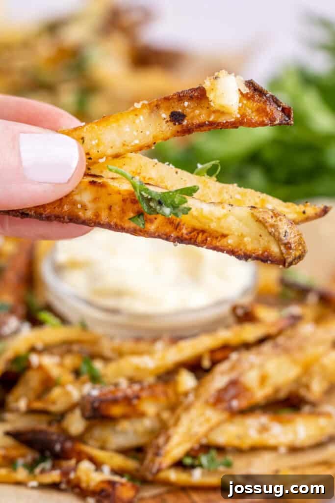 A hand reaching for a few crispy garlic parmesan fries, held over a plate piled high with more fries, and a jar of garlic aioli visible in the background.
