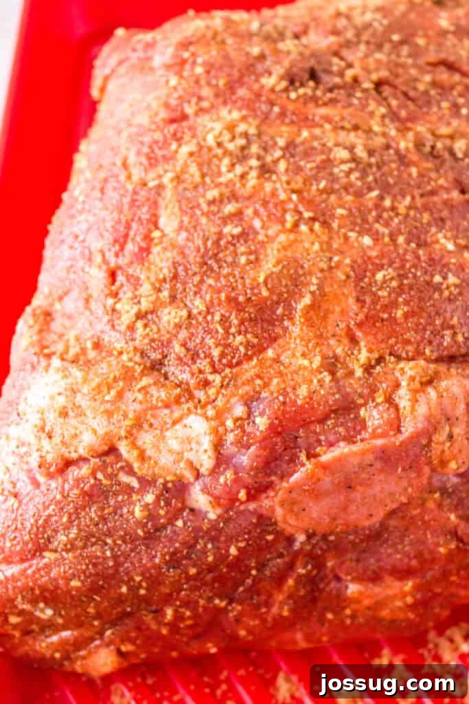 Raw Boston butt covered generously in dry rub, resting in a red tray, showing thorough seasoning.