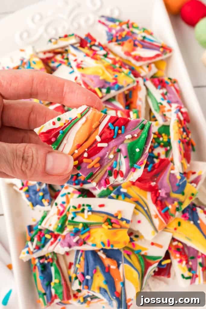 A hand gracefully holding a piece of homemade rainbow candy bark, showcasing its vibrant colors and intricate swirls, ready to be enjoyed as a delightful sweet treat.