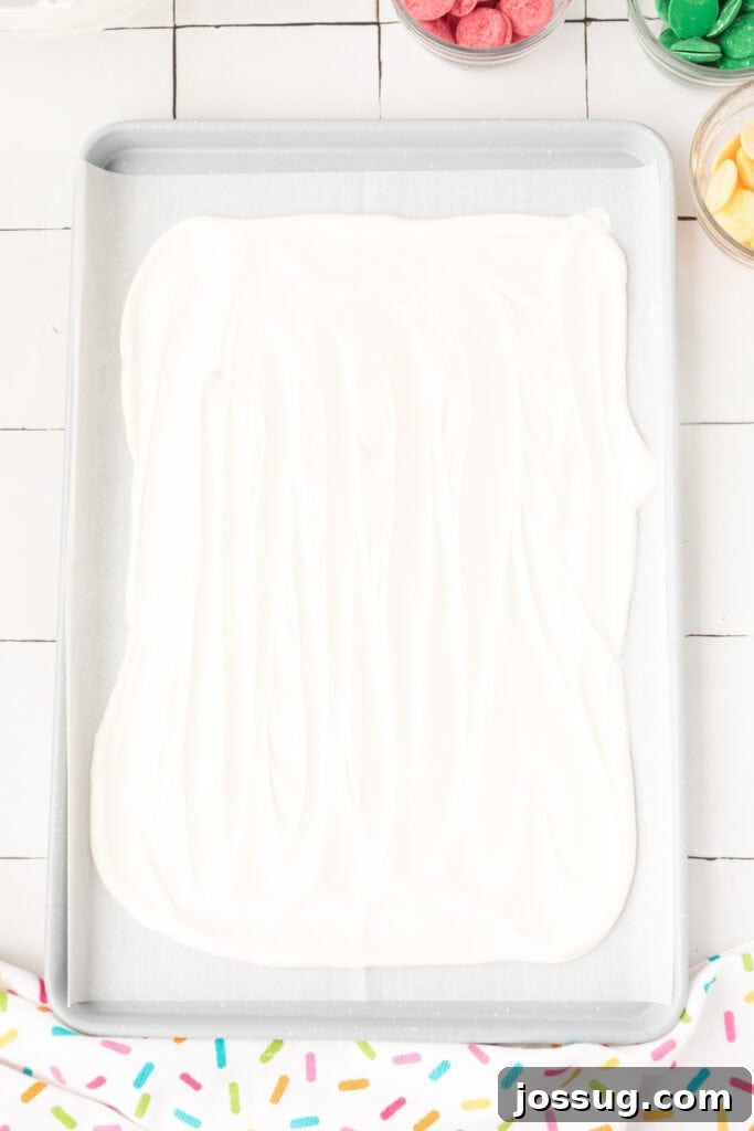 Melted white chocolate being poured and spread evenly onto a prepared baking sheet lined with parchment paper, forming a smooth, rectangular base for the rainbow candy bark.