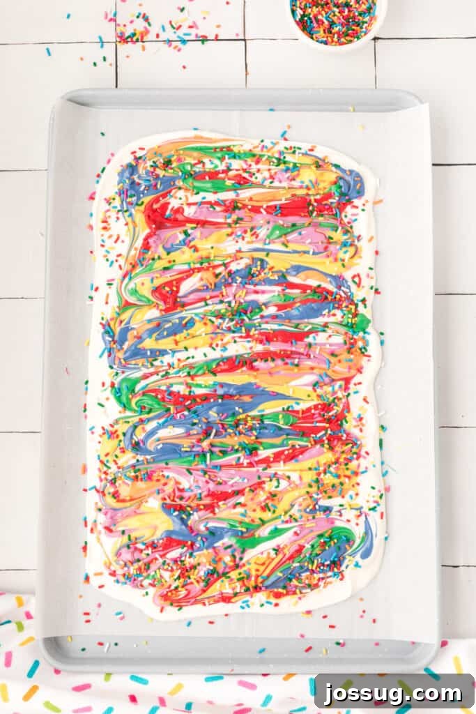 Finished rainbow candy bark, generously topped with colorful sprinkles, hardening on a baking sheet before being broken into delightful, edible pieces.
