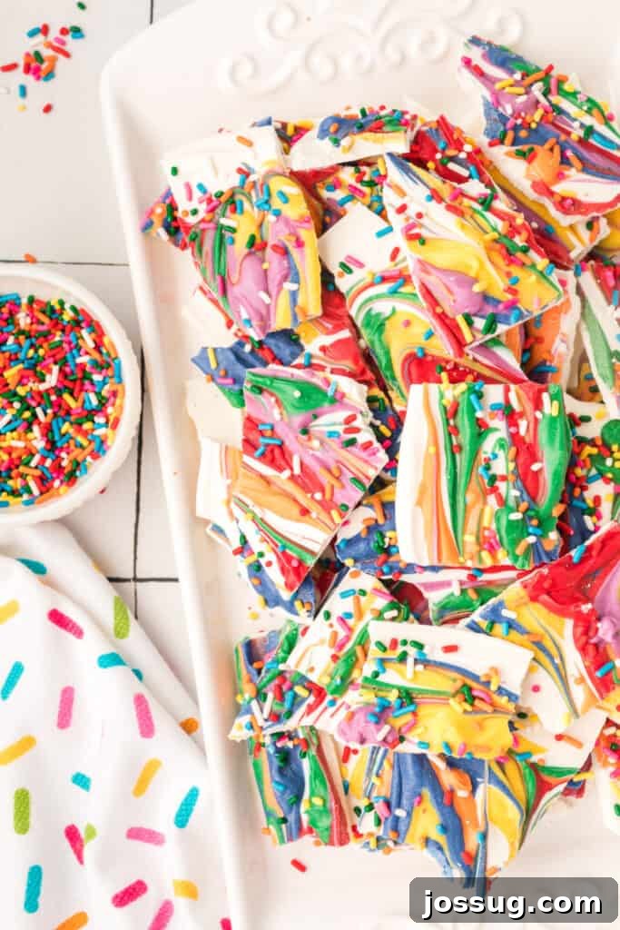 A vibrant plate of rainbow candy bark pieces, artfully arranged next to a small bowl filled with colorful sprinkles, ready for a festive celebration.