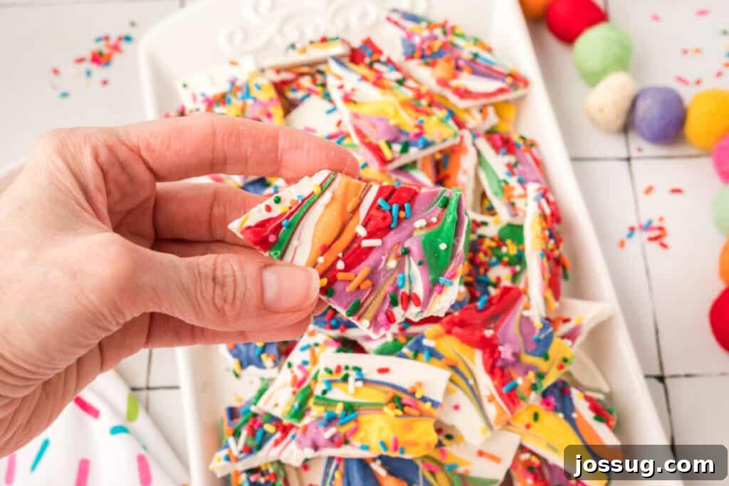 A close-up shot of a hand holding a piece of beautifully swirled rainbow candy bark, emphasizing the vibrant colors and appealing texture.