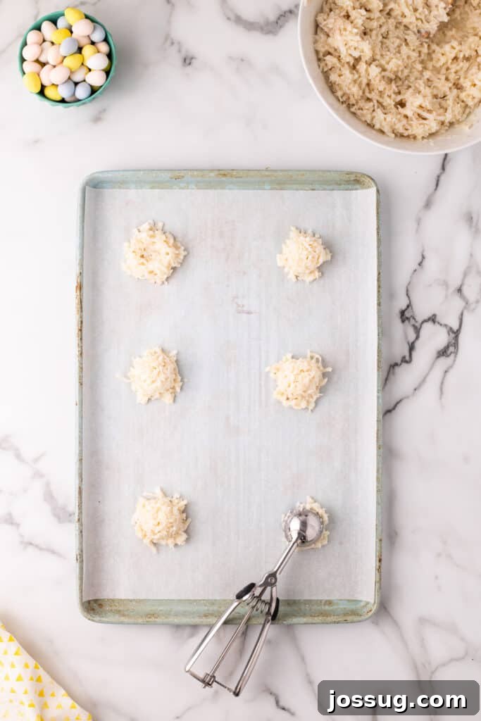 A cookie scoop places uniform mounds of coconut dough onto a parchment-lined baking sheet, preparing them for shaping into Easter nests.