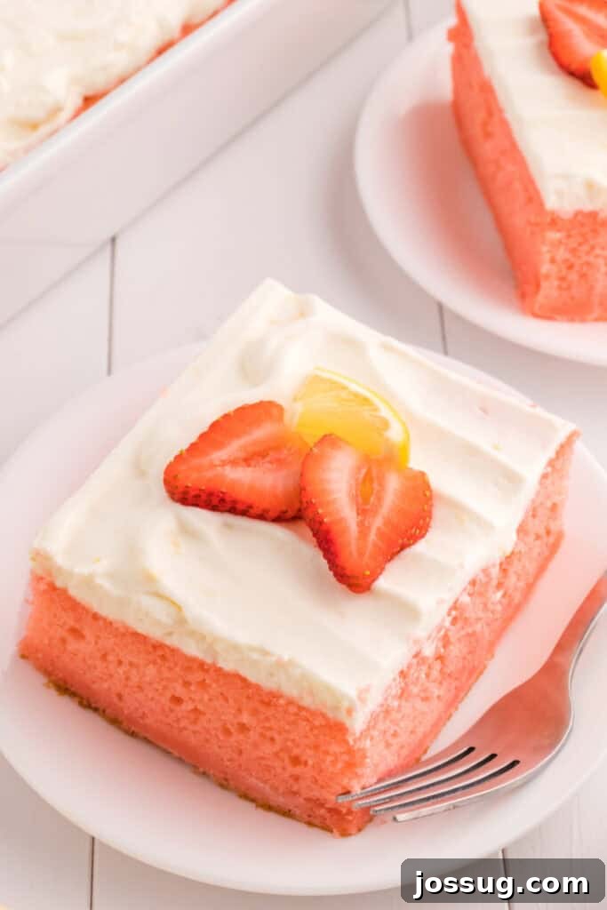 A top-down view of a generously frosted slice of strawberry cake with a light yellow lemon cream topping, garnished with fresh strawberry halves and thin lemon slices on a white plate.