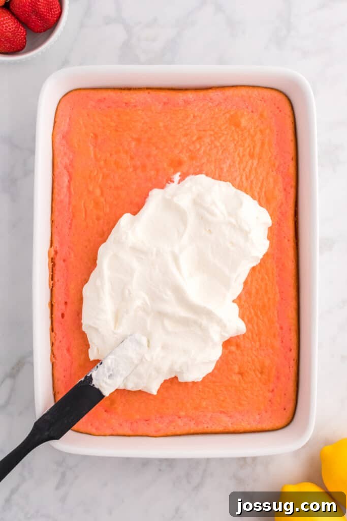 A spatula spreading the smooth lemon cream topping evenly over the surface of the cooled strawberry cake in the baking dish.