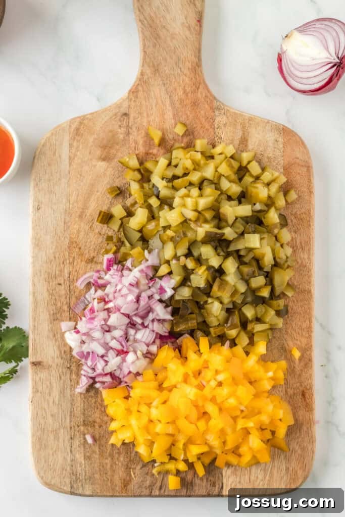Chopped dill pickles, red onion, and yellow bell pepper on a wooden cutting board, ready for mixing.
