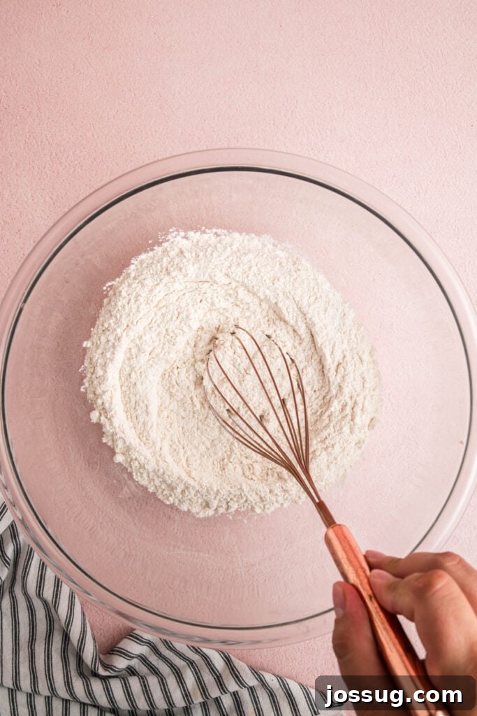 Sweet Strawberry Loaf 7 A hand using a metal whisk to thoroughly combine the dry ingredients in a clear glass mixing bowl, ensuring even distribution.